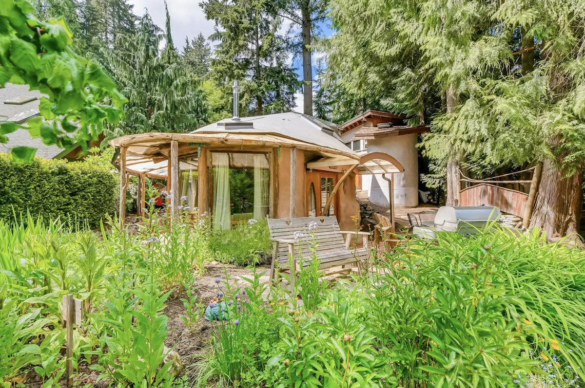 A hand-sculpted cob cottage with a rounded roofline, set behind a lush summer garden and ringed by tall conifers.