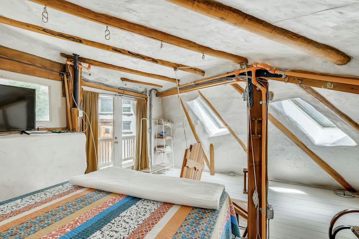 A loft bedroom under a hand-plastered white ceiling with exposed log beams, skylights, and a striped quilt.