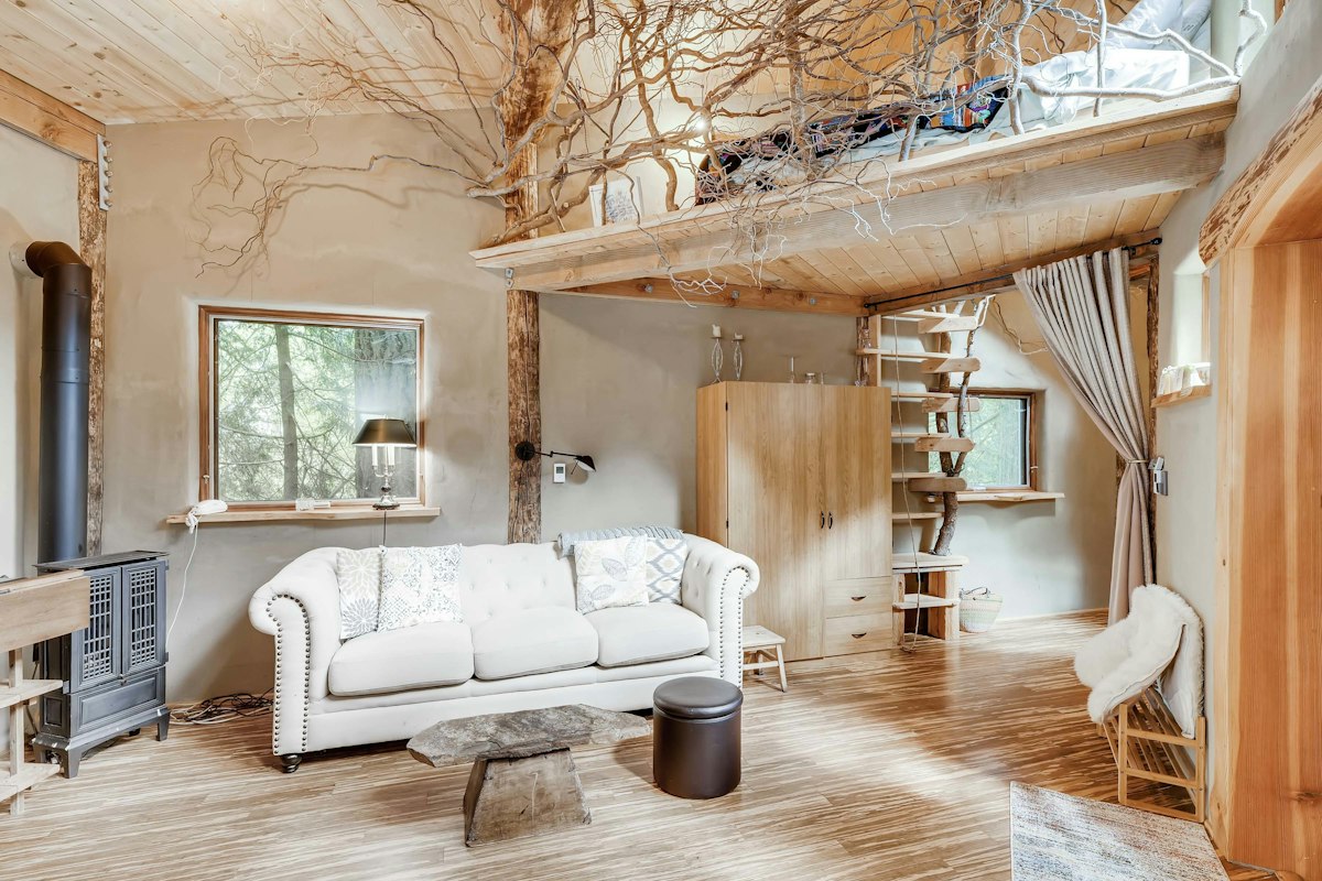 The living room of a cob cottage: a wood stove, a branch-relief loft overhead, a chesterfield sofa, and honey-wood floors.