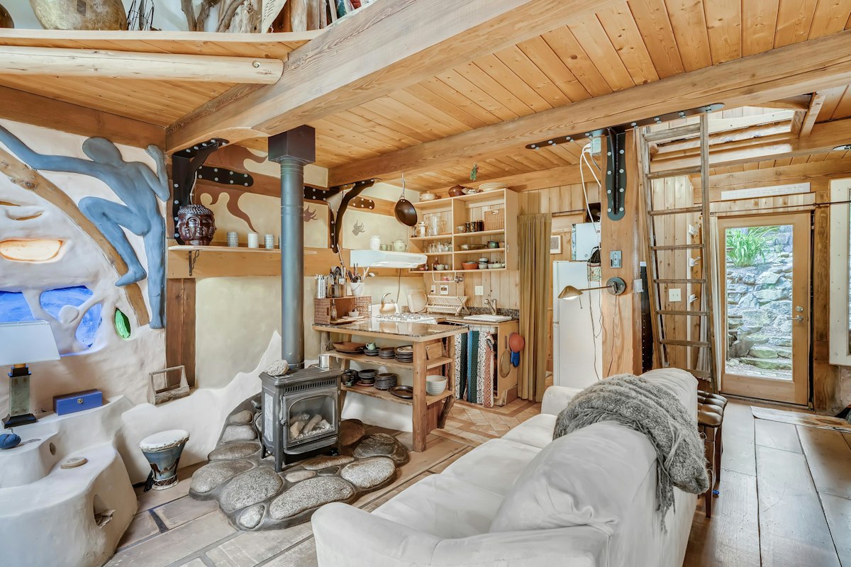 Inside the straw-bale studio: a hand-sculpted blue figure pressed into the plaster wall, a river-stone hearth around a wood stove, and a honey-wood ceiling.